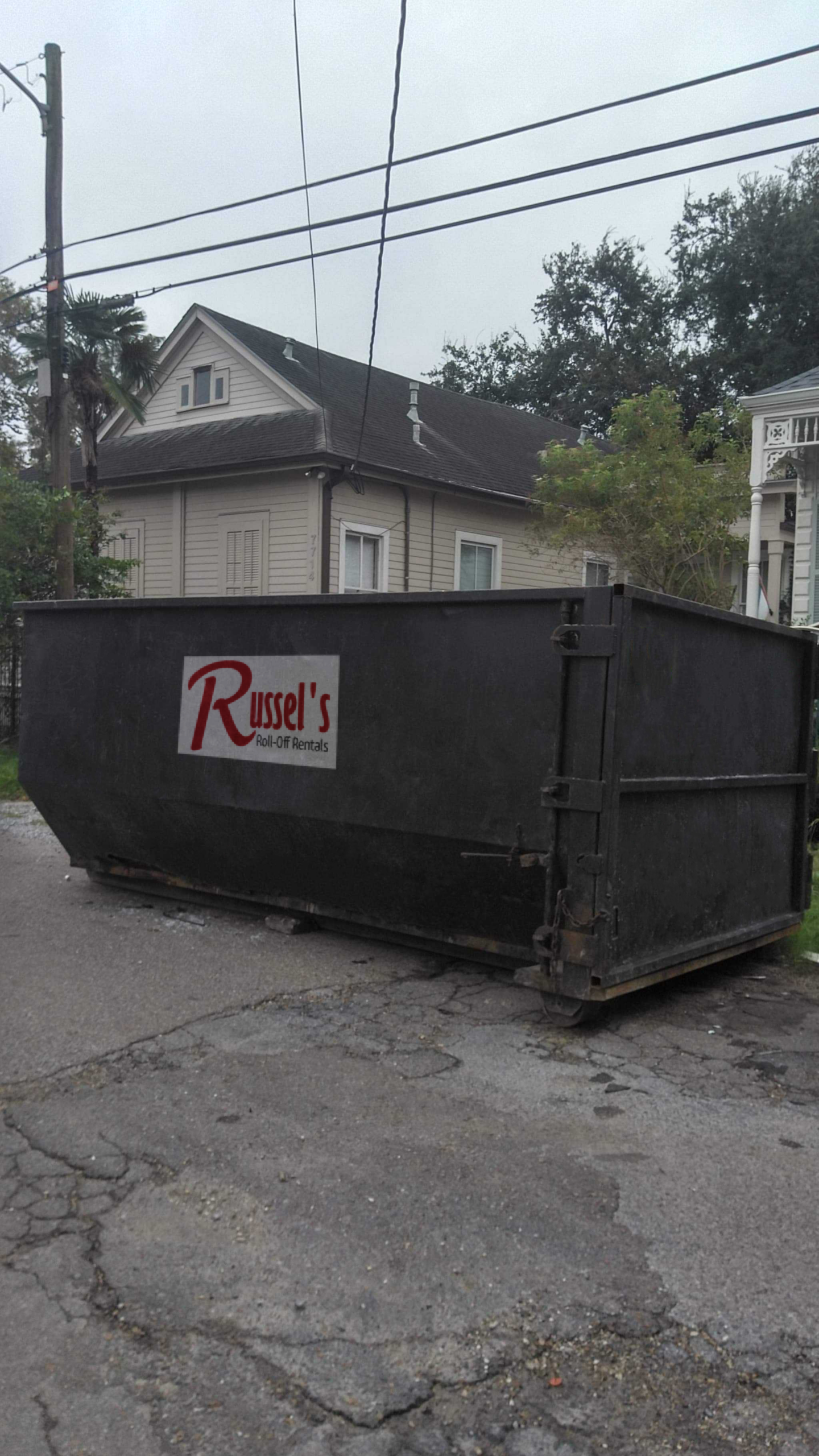 A large black dumpster with "Russel's Roll-Off Rentals" on the side sits on a cracked residential street. A house and trees are visible in the background.
